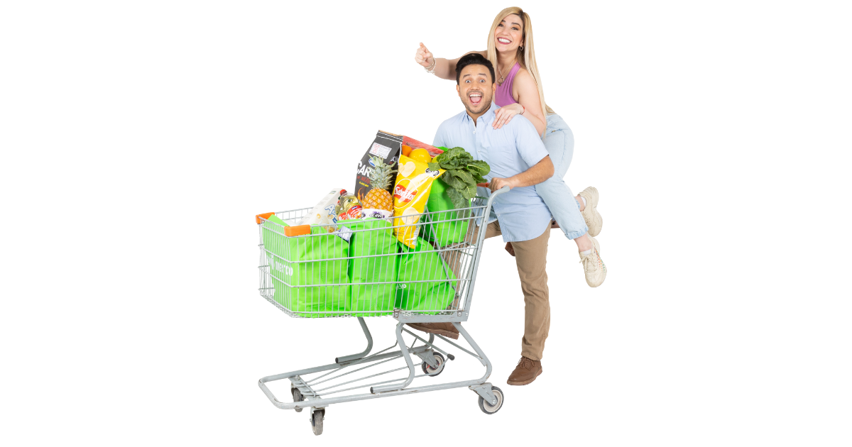Pareja sonriente junto a un carrito de supermercado lleno de productos Merco y bolsas verdes reutilizables.