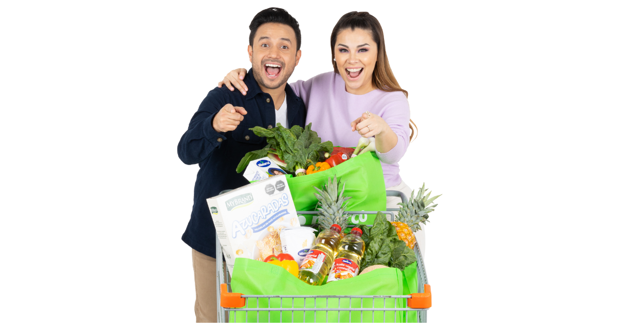 Pareja sonriente señalando hacia el frente mientras empuja un carrito de supermercado lleno de productos y bolsas verdes de Merco.